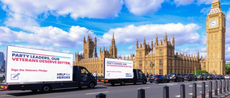 Vans with digi displays saying 'Party leaders, our veterans deserve better' drive in front of the Houses of Parliament